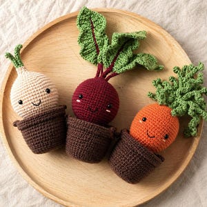 May include: Three crocheted vegetables: a white onion, a red beet, and an orange carrot, each with smiling faces and green tops, are displayed in brown pots on a wooden tray. The vegetables are handmade and have a whimsical, playful design.