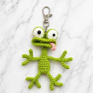 May include: A green crocheted frog keychain with large white-rimmed eyes and a pink tongue. The frog has outstretched arms and legs. The keychain has a silver-colored clasp.
