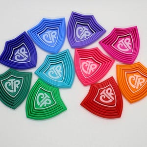 May include: A collection of colorful shield-shaped objects, each with a layered design and the letters "CTR" in the center. The shields are in various colors, including blue, green, pink, and orange, and are arranged on a white surface.