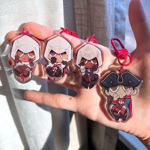 May include: Four clear acrylic keychains featuring cartoon characters. Three keychains depict a character in a white hood and red and brown clothing. The fourth keychain shows a character in a blue coat and black hat. All keychains have red lanyards.