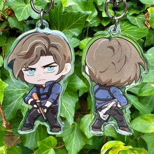 May include: Two clear acrylic keychains featuring a cartoon character in a blue uniform, black pants, and brown hair. One keychain shows the front view, the other the back. Both have silver key rings.