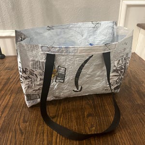 Könnte beinhalten: Eine wiederverwendbare Einkaufstasche aus recycelten Materialien. Die Tasche ist überwiegend silberfarben mit schwarzen Akzenten und hat einen schwarzen Griff. Das Amazon-Logo ist auf der Vorderseite der Tasche zu sehen. Die Tasche ist offen und bereit zum Befüllen.