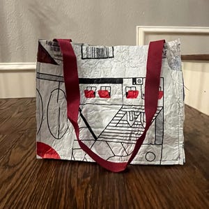Peut inclure: Sac fourre-tout rectangulaire avec un fond blanc et un motif graphique noir et rouge. Le sac a deux sangles bordeaux et est posé sur une surface en bois. Le motif comprend des formes géométriques et une maison.