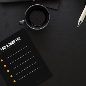 May include: A black notepad with the text "THE 'I DID A THING' LIST" and a series of lines and yellow dots. A black mug with coffee, a pen, and wireless earbuds are also on the black surface.