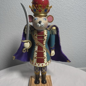 Puede incluir: Un cascanueces decorativo de madera con forma de rey ratón, sosteniendo una espada. El ratón lleva una corona roja y dorada, una chaqueta azul y roja y una capa morada. La base dice "El Cascanueces Rey Ratón".