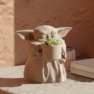 Puede incluir: Una maceta Grogu de color beige que sostiene una pequeña maceta cilíndrica con una suculenta verde. La figura tiene orejas grandes y está sentada. La maceta está sobre una superficie de color claro con un fondo borroso.