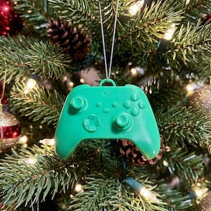 May include: A bright green Xbox controller Christmas tree ornament. The ornament is hanging from a silver string and is surrounded by a decorated Christmas tree with pine needles, ornaments, and lights. The controller has buttons and joysticks.