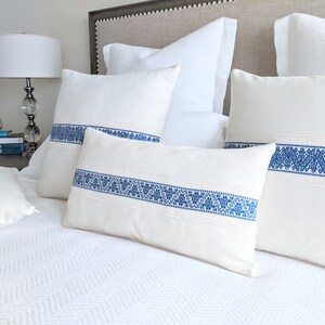 May include: Four white throw pillows with blue embroidered trim. The pillows are arranged on a white bed with a white headboard.