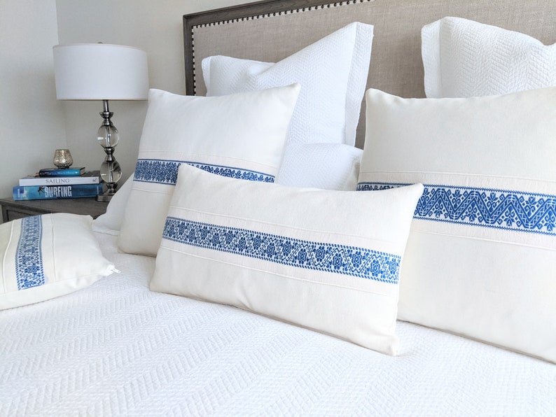 May include: Four white throw pillows with blue embroidered trim. The pillows are arranged on a white bed with a white headboard.