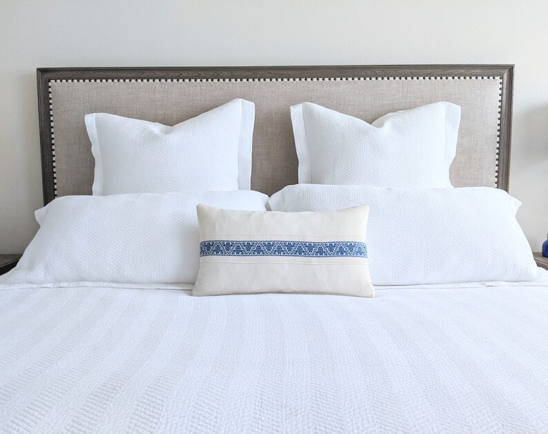 May include: A white bed with a gray headboard and white bedding. The bed has two white pillows and a decorative pillow with a blue stripe.