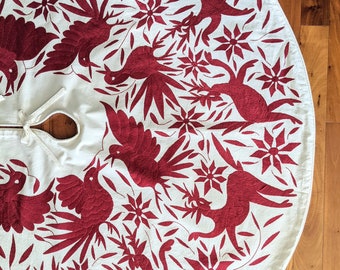 Pode incluir: Uma saia de árvore de algodão branco com um design bordado vermelho com pássaros e flores estilizados. A saia é circular e tem uma borda franjada.