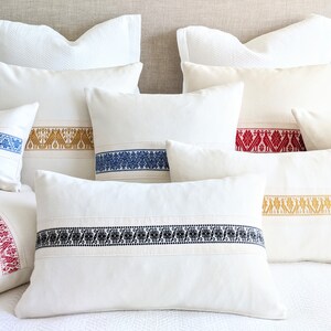 May include: A set of six white throw pillows with embroidered trim in various colors, including red, blue, gold, and black. The pillows are arranged on a white bed with a beige headboard.