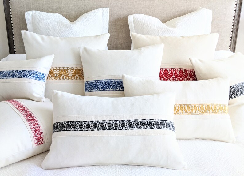 May include: A set of six white throw pillows with embroidered trim in various colors, including red, blue, gold, and black. The pillows are arranged on a white bed with a beige headboard.