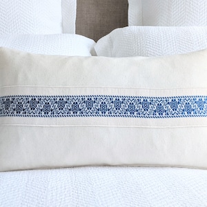 May include: A white linen pillow with a blue embroidered band across the center. The band features a repeating geometric pattern.