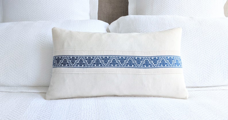 May include: A white linen pillow with a blue embroidered band across the center. The band features a repeating geometric pattern.