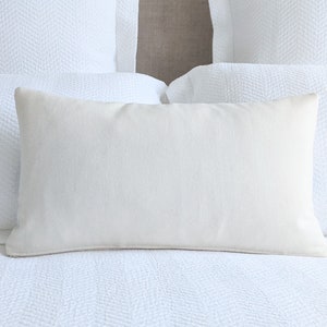 May include: A white rectangular pillow with a smooth, linen-like texture sits on a white bed with a herringbone pattern.