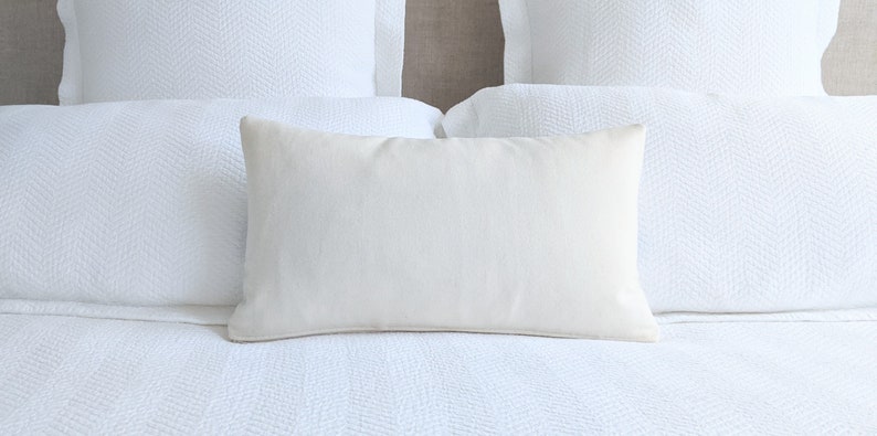 May include: A white rectangular pillow with a smooth, linen-like texture sits on a white bed with a herringbone pattern.