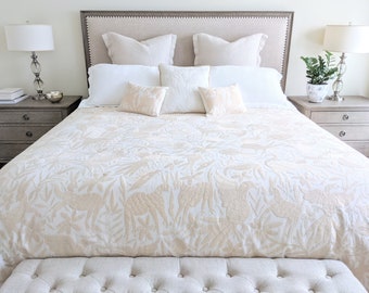 May include: A white bed with a beige embroidered quilt featuring a floral and animal design. The bed has a beige headboard and a white linen duvet cover. The bed is made up with white pillows and a beige throw pillow. The bed is sitting on a beige upholstered bench.