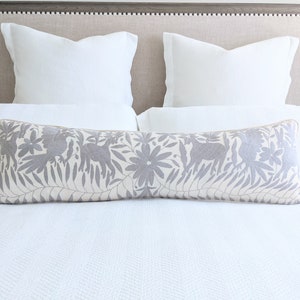 Pode incluir: Uma cama branca com um travesseiro lombar estampado em cinza e branco. A cama tem uma cabeceira cinza com capitonê branco.
