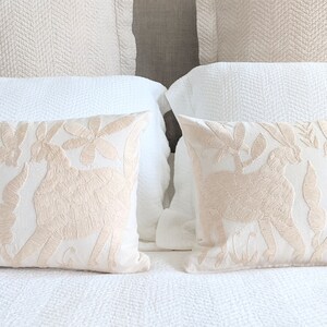 May include: Two embroidered throw pillows with a cream background and beige floral and animal designs. The pillows are on a white bed with a white and beige patterned headboard.