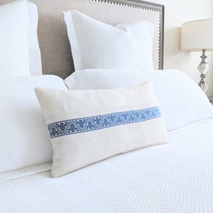 May include: A white bed with a white duvet cover and a white pillow with a blue embroidered band. The bed is made up with a white pillow and a white throw pillow with a blue embroidered band.