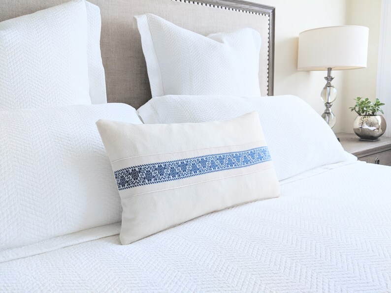 May include: A white bed with a white duvet cover and a white pillow with a blue embroidered band. The bed is made up with a white pillow and a white throw pillow with a blue embroidered band.