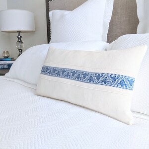 May include: A white pillow with a blue embroidered band across the center. The pillow is on a white bed with a white headboard.