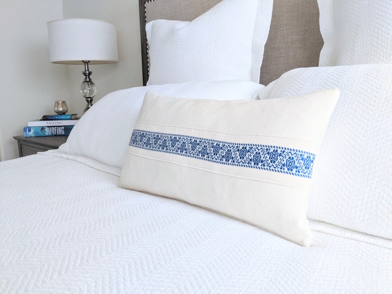 May include: A white pillow with a blue embroidered band across the center. The pillow is on a white bed with a white headboard.