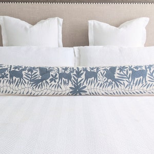 May include: A white bed with a white duvet cover and a blue and white embroidered bolster pillow. The pillow has a pattern of birds, flowers, and leaves.