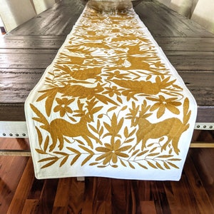 May include: A white table runner with a gold embroidered design of stylised birds and flowers. The runner is laid on a dark wood table.