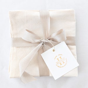 May include: A set of four off-white linen napkins tied with a beige ribbon and a gift tag. The tag has a gold logo that says "Sindo Huns, Handmade in Mexico by Otomi Textile".