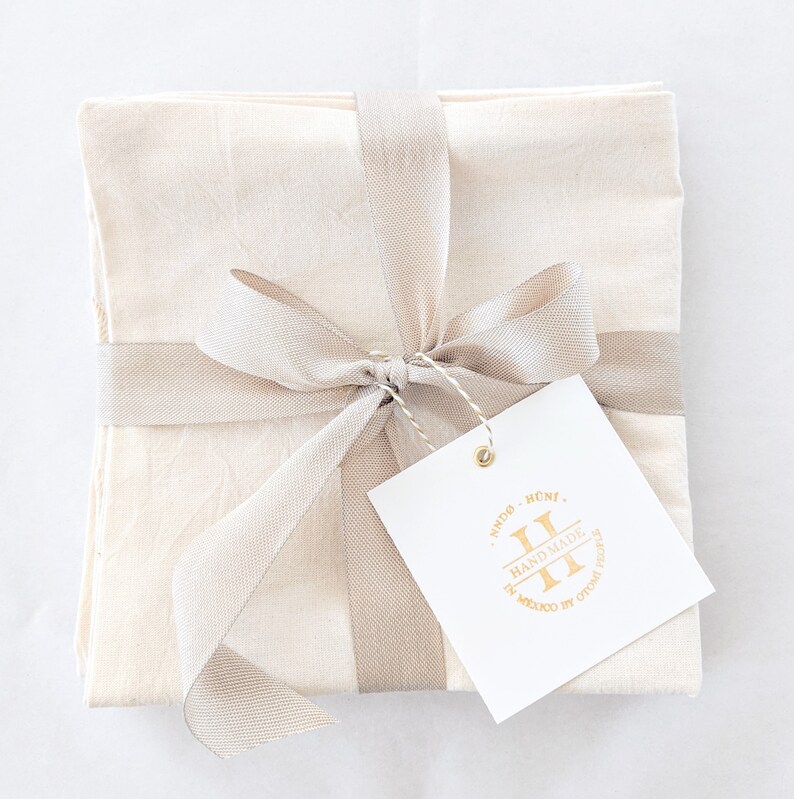 May include: A set of four off-white linen napkins tied with a beige ribbon and a gift tag. The tag has a gold logo that says "Sindo Huns, Handmade in Mexico by Otomi Textile".