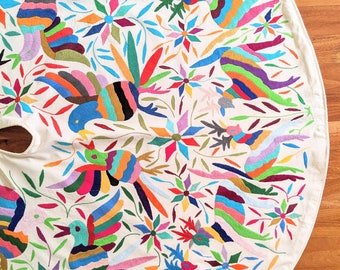 Pode incluir: Uma toalha de mesa circular branca com pássaros e flores bordados coloridos. O bordado é feito em estilo mexicano tradicional.