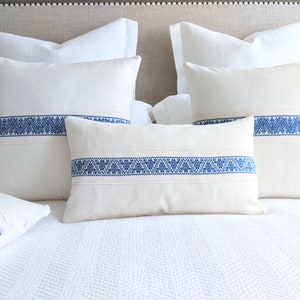 May include: Four white throw pillows with blue embroidered trim. The pillows are arranged on a white bed with a white headboard.