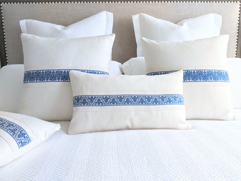 May include: Four white throw pillows with blue embroidered trim. The pillows are arranged on a white bed with a white headboard.