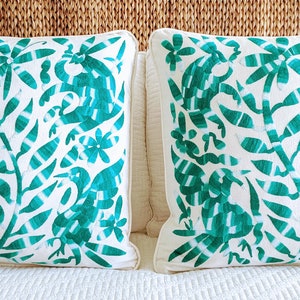 Op de afbeelding: Twee witte kussenslopen met een turquoise blauw geborduurd ontwerp van gestileerde vogels en bloemen. De kussens liggen op een wit beddengoed.