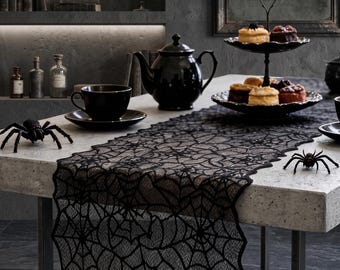May include: A black lace table runner featuring an intricate spider web pattern with scalloped edges. The runner is displayed on a light-coloured table alongside a black teapot, a matching teacup, a two-tiered serving tray with pastries, and two black spider figurines.
