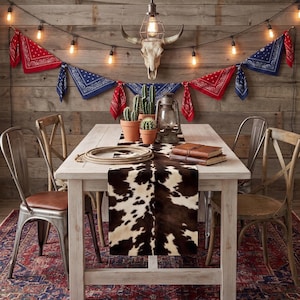 May include: A white wooden dining table featuring a cowhide print table runner, a coiled rope, two leather-bound journals, and two terracotta pots with green cacti. A metal lantern sits on the table, while a steer skull hangs on the wall above.