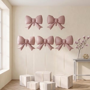 May include: Five dusty rose bow-shaped balloons are suspended in the air, each attached to a thin ribbon. Below the balloons, five white gift boxes of varying sizes are arranged on the floor, creating a coordinated decorative display.