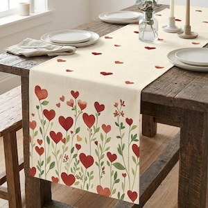 May include: A cream-colored table runner with a floral heart design. Red hearts of varying sizes are arranged on green stems and leaves. Scattered red hearts are also printed on the runner's surface. The runner is placed on a rustic wooden table.