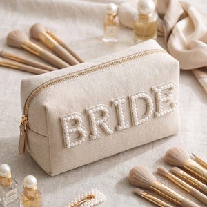 May include: A beige canvas makeup bag with the word "BRIDE" spelled out in pearl-like beads. The bag has a gold zipper and is surrounded by makeup brushes and perfume bottles.