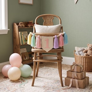 May include: A wooden high chair with a banner that says "1" in pink, decorated with pastel-colored tassels. The chair is set up for a first birthday celebration, with balloons and wrapped gifts nearby. A wooden bookshelf and a basket with a teddy bear are in the background.