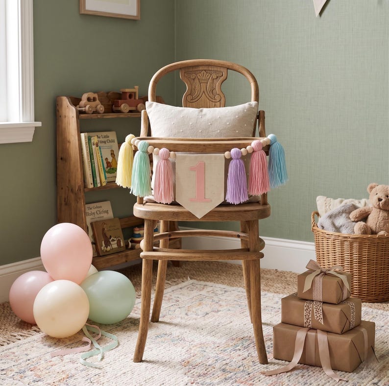 May include: A wooden high chair with a banner that says "1" in pink, decorated with pastel-colored tassels. The chair is set up for a first birthday celebration, with balloons and wrapped gifts nearby. A wooden bookshelf and a basket with a teddy bear are in the background.