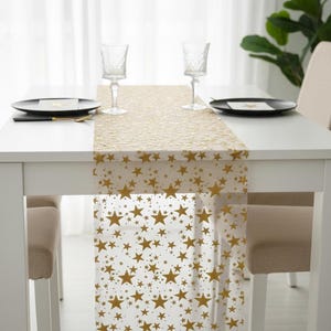 May include: A white table set for a meal with a gold star-patterned table runner. Two black plates, two wine glasses, and two chairs are visible. The table runner is made of a sheer material.