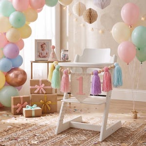 May include: A white high chair decorated for a first birthday celebration. Pastel balloons, gift boxes, and a banner with the number "1" and colorful tassels create a festive atmosphere. A framed photo and party hat are also visible.