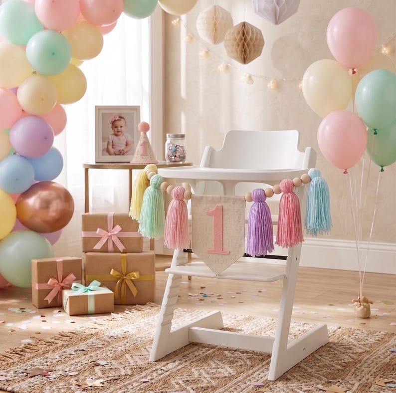 May include: A white high chair decorated for a first birthday celebration. Pastel balloons, gift boxes, and a banner with the number "1" and colorful tassels create a festive atmosphere. A framed photo and party hat are also visible.