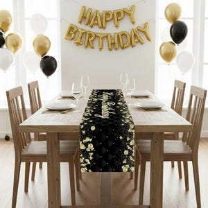 May include: A wooden dining table set for a celebration, featuring a black and gold table runner with a diamond pattern and gold floral accents. Gold balloons and a "Happy Birthday" banner complete the festive scene.