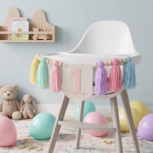 May include: A high chair decorated for a first birthday celebration. A banner with the number "1" and colorful tassels hangs across the front. Balloons in pastel colors and stuffed animals are in the background.
