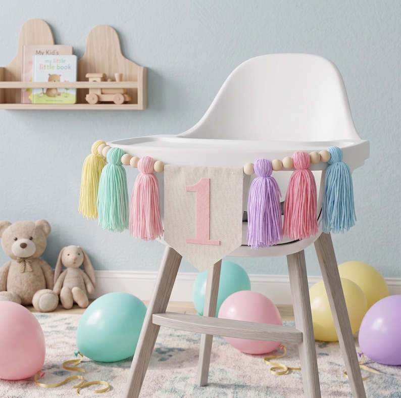 May include: A high chair decorated for a first birthday celebration. A banner with the number "1" and colorful tassels hangs across the front. Balloons in pastel colors and stuffed animals are in the background.