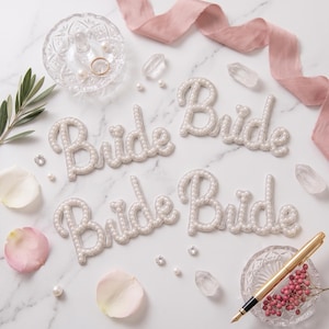 May include: Four white beaded "Bride" word cutouts are arranged on a marble surface, alongside a gold pen, ring, and decorative elements. The letters are outlined with small beads. Pink ribbon and rose petals add a touch of color.